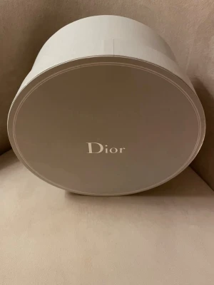 Rund förvaringsbox från Dior - Lyxig rund förvaringsbox från Dior i ljusgrå  kartong med elegant silvrig logga på locket. Perfekt för att förvara accessoarer eller använda som snygg inredningsdetalj. Stilren och exklusiv design som ger en lyxig känsla till rummet. Inköpt i Paris för ett par år sedan. Oanvänd så i mycket gott skick.