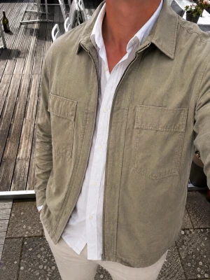 Zara Man Overshirt – Olivgrön/Khaki – Relaxed Fit - Snygg och stilren overshirt från Zara Man i en olivgrön/khaki nyans. Modellen har relaxed fit, dragkedja framtill, klassisk skjortkrage och två stora bröstfickor med detaljerad sömnad. Perfekt som ett lager över skjorta eller T-shirt och passar både till jeans och chinos.  Storlek: S Passform: Relaxed fit Färg: Olivgrön/khaki Märke: Zara Man Tillverkad i Turkiet