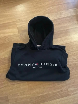 Svart hoodie från Tommy Hilfiger - Snygg svart hoodie från Tommy Hilfiger med klassisk logga och texten 'EST. 1985' på bröstet. Hoodien har huva med dragsko och en stor magficka. Perfekt för en avslappnad och stilren look. Saknar dock snören men annars fin