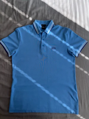 Hugo Boss pike - Snygg ljusblå pikétröja från BOSS med klassisk krage och knappar framtill. Tröjan har vita och mörkblå detaljer på kragen och ärmarna samt broderad BOSS-logga på bröstet. Perfekt till både jeans och shorts för en clean look.
