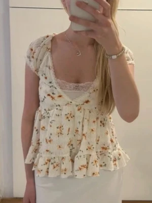 Blommig volangblus med v-ringning - Supersöt blus i ljus färg med små blommor i orange och grönt. Toppen har korta ärmar, v-ringning med spetskant och volang nertill. Materialet är ribbad bomull som ger en mjuk och stretchig känsla. Perfekt för vår och sommar!