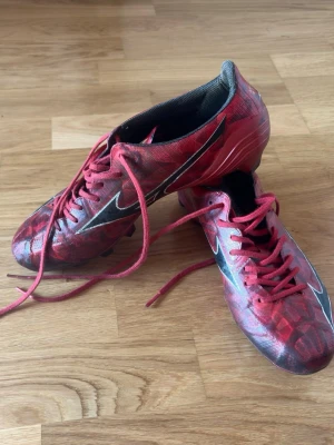 Röda Fotbollskor Mizuno a II Elite 40.5 - Mizuno fotbollsskor som köptes under Gothia Cup som en nödlösning efter att mina tidigare skor gick sönder. Skorna är hela och har inga skador eller defekter. De har endast använts under en kortare period. Legat på hyllan nu ett tag  Utseendet är något påverkat av användning på konstgräs, vilket kan synas på bland annat materialet lite svarta. I övrigt är skorna i fint och fullt fungerande skick.