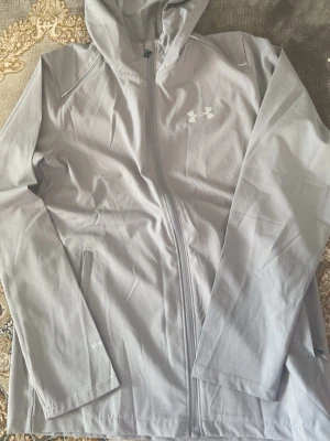 Ljusgrå vindjacka från Under Armour - Snygg ljusgrå vindjacka från Under Armour med huva och hel dragkedja framtill. Jackan har två sidofickor med dragkedja och logga på bröstet samt stor logotyp på ryggen. Tillverkad i lätt och vindtätt material, perfekt för blåsiga dagar.