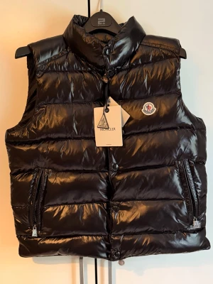 Svart dunväst från Moncler - Snygg svart dunväst från Moncler med glansig finish och hög krage. Västen har två dragkedjeförsedda fickor och klassisk Moncler-logga på bröstet. Perfekt för lager-på-lager och streetstyle. 
