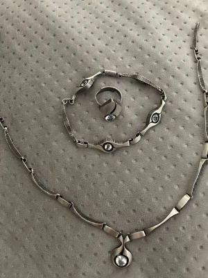 Smyckesset i silver och med bergkristaller - Snyggt smyckesset med halsband, armband och ring i silverfärgad. Alla delar har moderna, geometriska former och dekoreras med klara bergkristaller som ger en elegant touch. Perfekt för dig som gillar stilrena accessoarer med lite edge. De är från märke Karl Laine.