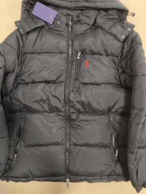 Svart pufferjacka från Ralph lauren - Snygg svart pufferjacka från Polo Ralph Lauren med röd broderad logga på bröstet. Jackan har dragkedja framtill, två sidofickor med dragkedja och en huva. Tillverkad i glansigt, vadderat material som håller dig varm under kalla dagar.
