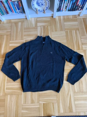 Mörkblå stickad tröja med halvzip - Mörkblå stickad tröja med halv dragkedja och ståkrage. Tröjan har långa ärmar, ribbade muddar och nederkant samt en diskret broderad logga på bröstet. Perfekt för lager-på-lager och enkel att matcha med jeans eller chinos.