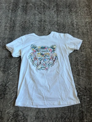 Vit Kenzo t-shirt med tigertryck - Säljer en vit t-shirt från Kenzo med det ikoniska tigertrycket i flera färger på bröstet. T-shirten har korta ärmar och en klassisk passform. Perfekt för dig som vill ha en statement piece i garderoben.