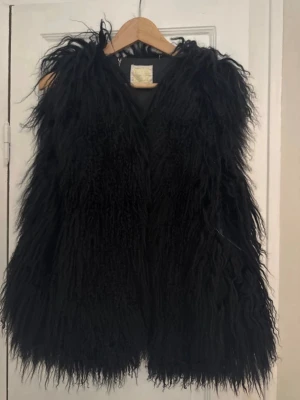 Svart långhårig pälsväst - Säljer en svart långhårig pälsväst med fluffig och voluminös look. Västen har en djup V-ringning och är ärmlös, perfekt för att ge din outfit en extra edge. Materialet är mongolian fur. Liten slitning på ena sidan. One size, passar xs-m