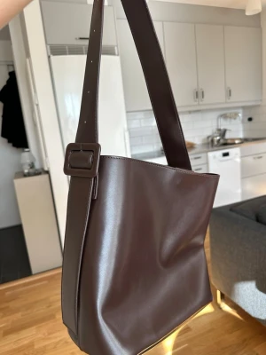 Brun väska  - Snygg brun axelväska i slätt konstläder med justerbart axelband och enkel, stilren design. Väskan har en fyrkantig form och stängs med magnetknapp. 