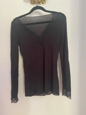 Svart långärmad topp med spetsdetaljer - Säljer en svart långärmad topp med v-ringning och spetsdetaljer vid halsringning och ärmslut. Tunn och stretchig modell som sitter snyggt på kroppen. Perfekt att styla med jeans eller kjol för en trendig look.