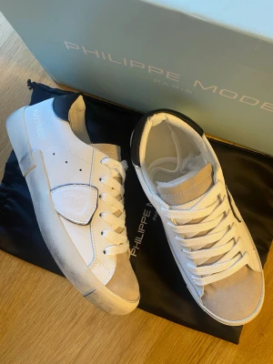 Vita Philippe Model sneakers med beige detaljer - Snygga vita sneakers från Philippe Model med beige mockadetaljer på tån och tungan. Skorna har vita snören, svarta detaljer vid hälen och en unik sidopanel med logotyp. Perfekta för dig som vill ha en stilren och trendig look.