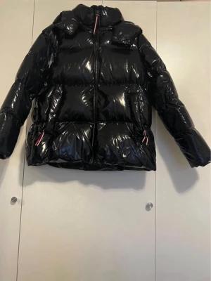Tommy hilfiger palmer glossy jacket - Säljer en riktigt snygg svart pufferjacka med glansig finish och hög krage. Jackan har dragkedja framtill och två fickor med detaljer i rött och vitt på dragkedjorna. Perfekt för kalla vinterdagar och ger en cool streetstyle-look.