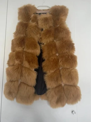 Brun pälsväst från Wonafa - Snygg brun pälsväst från Wonafa med fluffiga, fyrkantiga sektioner och svart innerfoder. Västen är ärmlös och har en modern, oversized look som ger en lyxig känsla. Perfekt för att lyfta din outfit med extra edge. 90 cm.