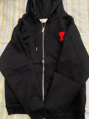 Svart hoodie med röd Ami-logo - Svart zip-hoodie från Ami med huva och dragsnören. På bröstet sitter Ami:s ikoniska röda hjärta med A-bokstav broderat. Hoodien har fickor framtill och silverfärgad dragkedja. Perfekt för en chill och stilren look.