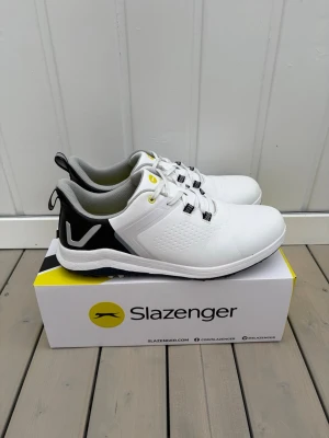 Vita golfskor från Slazenger - Snygga vita golfskor från Slazenger med svarta och gula detaljer. Skorna har en sportig design, grå insida och robust sula med greppvänliga dobbar. Perfekta för golfbanan och tillverkade i slitstarkt syntetmaterial. Endast använda 1 gång (fel storlek för mig). På kartongen står det stl 43, men det är stl 44. 
