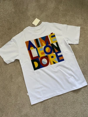 Vit Aimé Leon Dore t-shirt med färgglatt tryck - Säljer en vit t-shirt från Aimé Leon Dore med ett stort färgglatt grafiskt tryck på ryggen och ett mindre på bröstet. T-shirten har klassisk passform och är tillverkad i mjuk bomull. Perfekt för dig som gillar streetwear och snygga detaljer.