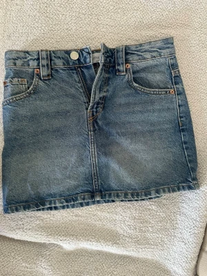 Blå jeanskjol med fem fickor - Säljer en klassisk blå jeanskjol i kort modell med fem fickor och bälteshällor. Kjolen har knapp och dragkedja framtill och är gjord i ett slitstarkt denimtyg. Perfekt att styla med sneakers eller boots. Aldrig använd pga för stor. Pris går att diskutera💗