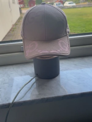 Ljusrosa keps med GG-mönster - Säljer en ljusrosa keps med böjd skärm och stort GG-mönster i samma ton över hela kepsen. Kepsen har klassisk form och är tillverkad i ett tygmaterial som känns slitstarkt och trendigt. Perfekt accessoar för att lyfta din outfit. Den är i lite dålig form där bak men inte så farligt och formen syns inte när man har på sig den