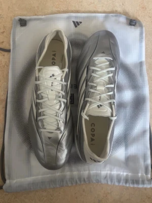 Adidas Copa fotbollsskor (använd på riksläger) - Snygga Adidas Copa fotbollsskor i silver och vitt med klassisk snörning. Skorna har en modern, strömlinjeformad design och är tillverkade i syntetmaterial med detaljer i läder. Perfekta för dig som vill sticka ut på planen med stilrena och sportiga skor.