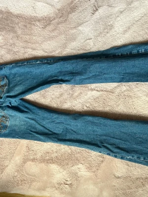Blå bootcut jeans med detaljer - Säljer ett par blå bootcut jeans med snygga slitningar och dekorativa detaljer på bakfickorna. Jeansen har klassisk femficksmodell och är tillverkade i denim med lätt stretch. Perfekta för dig som gillar en retroinspirerad look och vill ha ett par jeans som sticker ut. om ni behöver mått är det bara att skriva