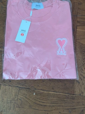 Rosa t-shirt från Ami med hjärtdetalj - Säljer en rosa t-shirt från Ami med ett broderat hjärta och A-logga på bröstet. T-shirten har rund hals och korta ärmar, perfekt för dig som gillar stilrena plagg med en twist.