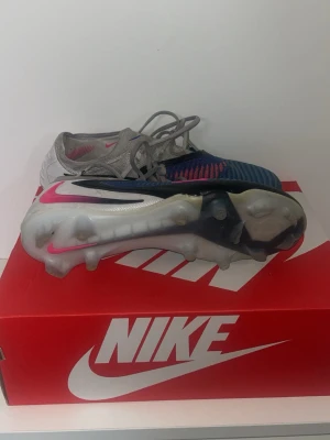 Nike blå och silver fotbollsskor - Säljer ett par Nike fotbollsskor i blått och silver med rosa och svarta detaljer. Skorna har snörning och en ovandel i textilmaterial som ger bra känsla på bollen. Sulan har fasta dobbar för spel på gräs. Snygg och modern design för dig som vill sticka ut på planen.