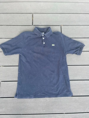Mörkblå pikétröja  - Klassisk mörkblå pikétröja i märket Lacoste med krage och tre knappar framtill. Tröjan har korta ärmar och en liten broderad logga på bröstet. Perfekt till en avslappnad stil och enkel att matcha med jeans eller shorts. Storlek XL