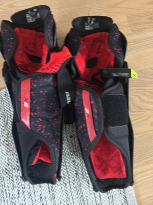 Bauer 3X benskydd hockey svart/röd - Säljer ett par Bauer 3X benskydd för hockey i svart och rött. Skydden har ventilerande hål, justerbara kardborreband och slitstark plast på framsidan. Insidan är vadderad för extra komfort och stötdämpning. Perfekt för dig som vill ha bra skydd på isen.