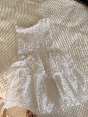 Vit kort klänning från RYVLS - Supersöt vit kort klänning från RYVLS med smock upptill och volangig kjol. Klänningen är ärmlös och har broderade spetsdetaljer som ger en romantisk känsla. Perfekt för varma dagar och somriga tillfällen.