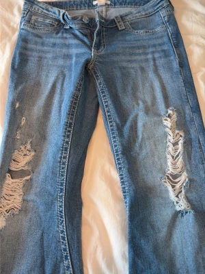 Blå jeans med hål - Säljer ett par blå jeans med stora slitningar och hål på båda benen. Jeansen har klassisk femficksmodell, knappgylf och dekorativa bakfickor med lock och knapp. 