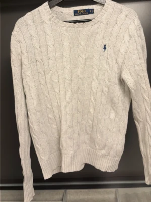 Vit kabelstickad tröja Polo Ralph Lauren - Vit kabelstickad tröja från Polo Ralph Lauren med rund halsringning och blå broderad logga på bröstet. Tröjan har ribbade muddar och är tillverkad i ett mjukt stickat material. Perfekt för en klassisk och stilren look. Har ett litet hål som man ser och lite slitet i kragen annars som ny
