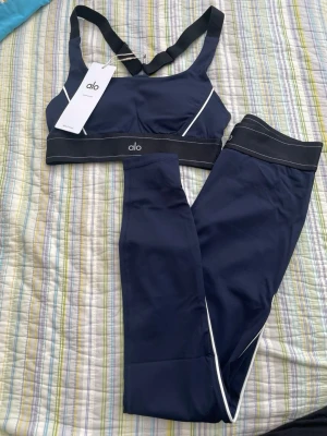 Mörkblått set från Alo med vita detaljer - Snyggt mörkblått set från Alo med sport-bh och matchande tights. Setet har vita kontrastdetaljer och breda svarta resårband med Alo-logga. Materialet är stretchigt och följsamt, perfekt för en aktiv livsstil.