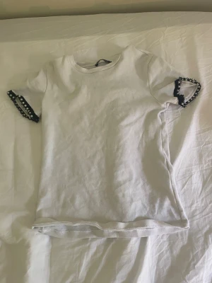 Vit t-shirt med spetsdetaljer - Säljer en vit t-shirt med rund hals och korta ärmar. Ärmsluten har svarta spetsdetaljer med små vita pärlor som ger en unik touch. T-shirten är i mjukt material och har en enkel, rak passform. I storlek S.