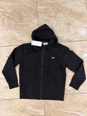 Svart zip hoodie från Lacoste - Säljer en svart hoodie från Lacoste med dragkedja framtill och klassisk krokodil-logga på bröstet. Hoodien har huva med dragsko, två stora fickor fram och ribbade muddar vid ärmar och midja. Tillverkad i mjukt bomullsmaterial för skön känsla.