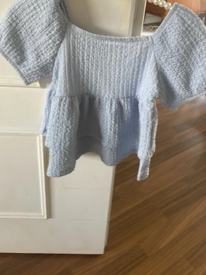 Ljusblå topp med puffärmar - Supersöt ljusblå topp med puffiga korta ärmar och strukturerat tyg. Toppen har en volang nedtill och en lös, flowy passform som ger en romantisk vibe. Perfekt att styla med jeans eller kjol för en trendig look.