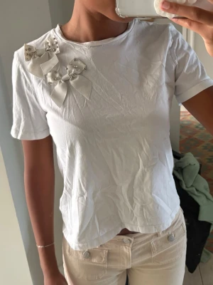 Vit t-shirt med rosetter och pärlor - Säljer en vit t-shirt med korta ärmar och rund hals. På axeln sitter två dekorativa rosetter med pärlor och små nitar som ger en söt och unik touch. Perfekt för dig som vill ha något extra till din outfit.
