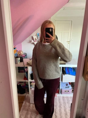 Grön stickad tröja med pärlor - Mysig grå stickad tröja med rund hals och dekorativa pärlor och små stenar på bröstet. Tröjan har en avslappnad passform och långa ärmar med ribbade muddar. Perfekt för dig som gillar en enkel men ändå lite extra look.