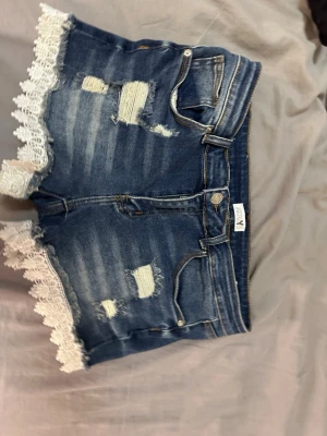 Blå jeansshorts med spetskant - Snygga blå jeansshorts med slitna detaljer och vit spetskant längs bensluten. Klassisk femficksmodell med dragkedja och knapp. Perfekta för en trendig och avslappnad sommarlook.