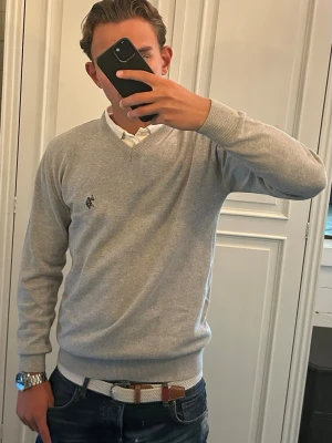 Ljusgrå stickad tröja U.S. Polo Assn. - Snygg ljusgrå stickad tröja från U.S. Polo Assn. med V-ringning och diskret logga broderad på bröstet. Tröjan har ribbade muddar vid ärmslut och nederkant, perfekt att bära över en skjorta för en clean och stilren look.