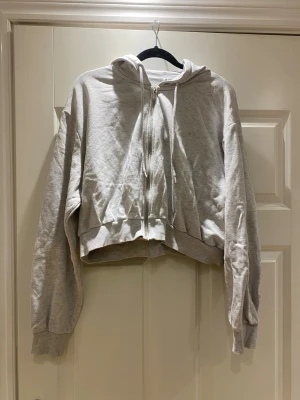 Ljusgrå croppad hoodie med dragkedja - Säljer en ljusgrå croppad hoodie med dragkedja framtill och huva med dragsnören. Hoodien har långa ärmar och bred mudd vid ärmslut och nederkant. Perfekt för en avslappnad och chill stil. Materialet känns mjukt och bekvämt, troligen bomullsblandning.