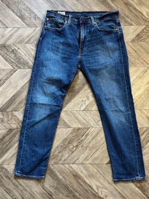 Levi's 502  - Säljer ett par klassiska blå jeans med rak passform och fem fickor. Jeansen har en djupblå tvätt med lätt slitning och kontrastsömmar. Gilfen är valdigt trög/fast Materialet är denim i bomull och de har bälteshällor samt knappgylf. Perfekta för en avslappnad stil. Längd 97cm Bredd 42cm
