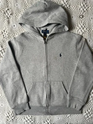 Grå zip hoodie från Polo Ralph Lauren - Klassisk grå hoodie med dragkedja från Polo Ralph Lauren. Tröjan har huva, två fickor framtill och den ikoniska mörkblå loggan broderad på bröstet. Tillverkad i mjuk bomullsblandning för en skön och avslappnad look.