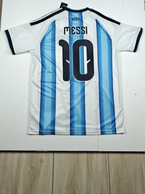 Argentina Messi matchtröja blåvit - Säljer en officiell Argentina matchtröja från Adidas med Messi och nummer 10 på ryggen. Tröjan har klassiska blå och vita vertikala ränder, svarta detaljer på axlar och ärmslut samt AFA-emblem och guldiga detaljer framtill. Perfekt för fotbollsfans!