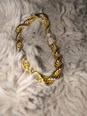 Guldtonat tvinnat armband 6mm - Snyggt armband i guldfärgad metall med tvinnad kedja och ett lås dekorerat med små gnistrande stenar. Armbandet har en chunky känsla och passar perfekt för dig som gillar statement-smycken och vill sticka ut lite extra.