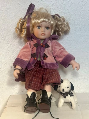 Porslinsdocka med lockigt hår och hund - Söt porslinsdocka med blont lockigt hår och lila rosetter. Dockan har en rosa och lila sammetsjacka, rutiga byxor och svarta kängor. Medföljer gör en liten vit dalmatinerhund i tyg. Perfekt för samlare eller dekoration.
