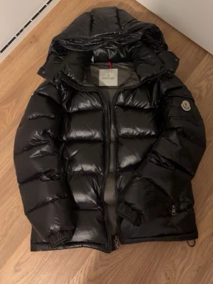 Svart dunjacka från Moncler - Snygg svart dunjacka från Moncler med glansig finish och klassisk pufferdesign. Jackan har huva, dragkedja framtill, innerficka med dragkedja och Moncler-logga på ärmen. Perfekt för kalla vinterdagar och riktigt varm.