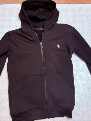 Svart hoodie från Polo Ralph Lauren - Svart hoodie med dragkedja från Polo Ralph Lauren. Klassisk design med vit broderad logga på bröstet, huva med snörning och känguruficka framtill. Tillverkad i mjukt bomullsmaterial som känns skönt mot huden.