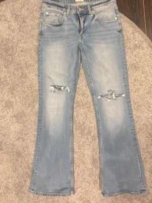 Ljusblå bootcut jeans med slitningar - Säljer ett par ljusblå bootcut jeans med slitningar på knäna och lätt tvättad look. Jeansen har fem fickor och klassisk knappgylf. Perfekta för dig som gillar en avslappnad och trendig stil.