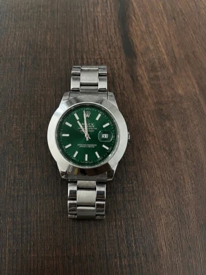 Justerbar silver klocka med grön urtavla - Snygg klocka i stål med länkarmband och grön urtavla. Klockan har tydliga indexmarkeringar, datumfönster och klassisk rund form. Perfekt accessoar för dig som vill sticka ut med stilren design. Den kommer med extra delar för att göra den större för personer som behöver det.